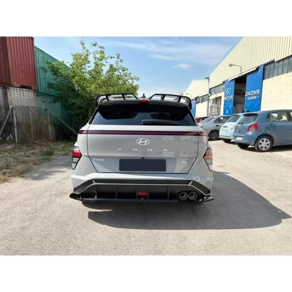 a full rear view of a light grey Hyundai Kona N-Line Mk2 parked on a sunlit road outside a workshop. Mounted at the lower edge of the rear bumper is a gloss black rear diffuser with multiple vertical bars (or fins) evenly spaced along its width. These bars protrude slightly downward, giving the vehicle a performance-oriented, aerodynamic appearance. The diffuser frames the dual exhaust outlets on the right side, enhancing the aggressive styling. It also includes a central cutout area—possibly for a red accent or faux tow hook—for added motorsport flair. The gloss finish reflects sunlight, contrasting with the matte textures of surrounding trim pieces.