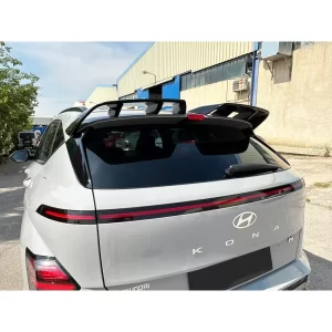 This image provides a closer look at the gloss black roof spoiler from the upper left rear of the vehicle. The spoiler is securely attached at both ends and arches over the rear brake light. Its high-gloss finish contrasts with the matte trim of the rear windshield and body paint. The shape and placement create a distinctive visual break above the rear hatch glass, reinforcing the Kona N-Line’s sporty, performance-driven styling.