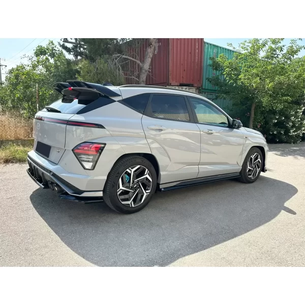 a light grey Hyundai Kona N Line from the rear-side angle, parked on an asphalt road with a semi-rural backdrop including trees and stacked shipping containers. The vehicle features gloss black side skirt extensions installed along the bottom edges of the side doors. These sleek, flat additions subtly enhance the sporty profile of the car, visually lowering its stance. The gloss black finish contrasts sharply against the body color, aligning with other black accents on the rear diffuser, spoiler, and wheels, contributing to a cohesive and aggressive look.