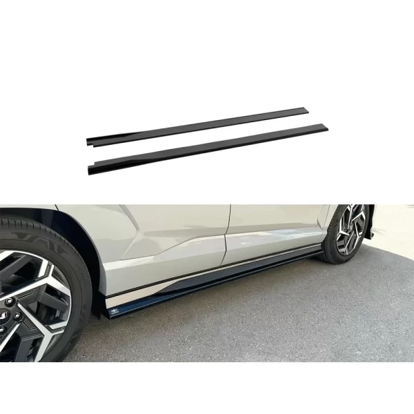 The image is divided into two parts. At the top, two gloss black side skirt extensions are shown individually against a white background. They are long, slim, and straight, designed to be mounted along the lower edges of the vehicle's body. Below, a close-up of the passenger side of the Hyundai Kona N Line shows the skirt extension installed. The part hugs the underside of the vehicle, sitting flush with the lower door panel. Its glossy black finish enhances the car's sporty aesthetic while offering subtle protection from road debris.
