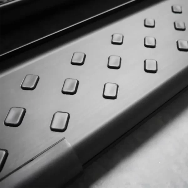 A detailed close-up of the walking surface of a black side step. The metal surface is matte with a slightly brushed finish and features evenly spaced, raised rectangular grips arranged in a grid pattern. These rubberized protrusions are designed to prevent slipping, especially in wet or muddy conditions.