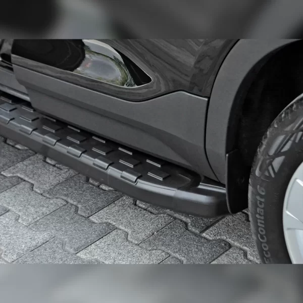 The image shows a close-up of a black SUV parked on grey interlocking stone pavement. Attached beneath the vehicle’s side door is a sturdy, matte-black side step. The step runs horizontally along the body and features a wide, flat surface with evenly spaced rectangular raised pads for anti-slip traction. The contour of the step curves to match the shape of the vehicle’s side skirt. It sits just below the door level, providing a stable platform to help passengers step in or out of the vehicle more safely—especially helpful for children, seniors, or individuals with mobility limitations.