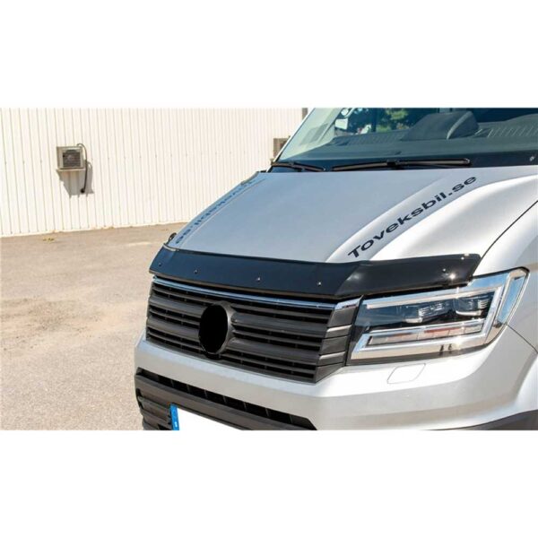 Glossy black hood deflector installed on the front edge of a silver van’s hood, stretching across its full width. It sits just above the vehicle’s grille and headlights, slightly raised with a gentle curve to divert airflow. The deflector has a smooth, shiny surface and is fastened with small visible bolts. The van features a wide front grille with horizontal slats and sharply contoured headlights with LED elements. The word "Toveksbil.se" is printed diagonally across the upper right part of the hood.