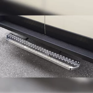 Sturdy rear step bar made of polished stainless steel, mounted just below the rear door of a commercial van. The step is rectangular with beveled corners and is covered with evenly spaced circular grip holes that provide slip resistance. The metallic surface is slightly reflective, showing a rugged, industrial look. Two black mounting brackets secure it firmly to the underside of the vehicle. The textured design allows safe footing when accessing the rear cargo area.
