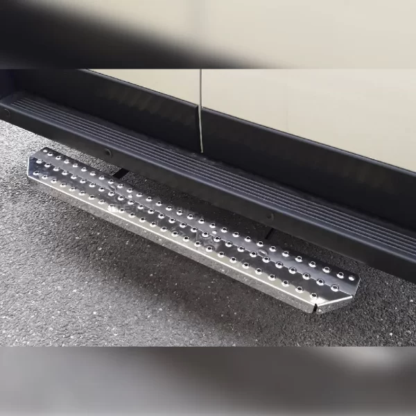 Sturdy rear step bar made of polished stainless steel, mounted just below the rear door of a commercial van. The step is rectangular with beveled corners and is covered with evenly spaced circular grip holes that provide slip resistance. The metallic surface is slightly reflective, showing a rugged, industrial look. Two black mounting brackets secure it firmly to the underside of the vehicle. The textured design allows safe footing when accessing the rear cargo area.